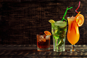 Cocktails with straws, ice and fruits on a black background. Alcoholic drinks on an old wooden...