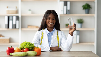 Young female african american nutritionist holding glass of water while sitting at table with...