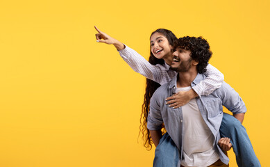 Excited young indian couple having fun and lady piggybacking guy and pointing finger at free space...
