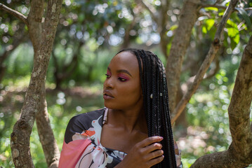 Portrait of Young African Woman standing under a tree in a mango orchard with long braids - obrazy, fototapety, plakaty