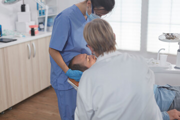 Fototapeta premium Clinical orthodontist team with face mask examining patient mouth during medical surgery in stomatological office room. Senior woman doctor using professional dentistry tools for cavity procedure
