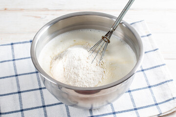 Cooking dough for traditional thin pancakes. Eggs, flour, salt, sugar, milk and butter in a bowl on a white wooden background. Recipe step by step. Food for Maslenitsa