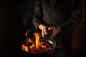 Chef prepares vegetable food in a frying pan with a flame of fire. The concept of restaurant and hotel service. Flamber or cooking over natural fire. Grande cuisine.