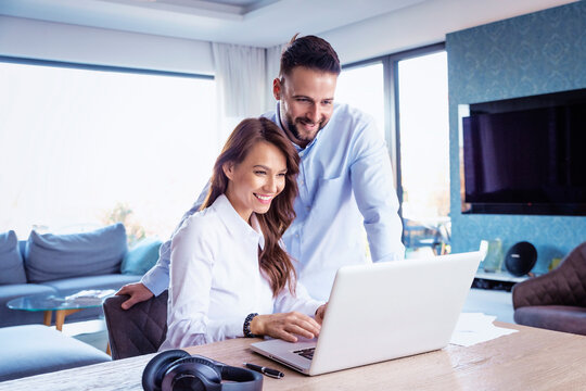 Smiling Woman Sitting At Desk Behind The Laptop While Handsome Man Standing Next To Her And Working Together