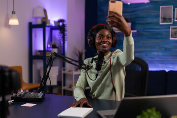Social media influencer holding smartphone taking selfie while recording vlog content using vlogging production equipment. African american vlogger woman hosting on air talk show
