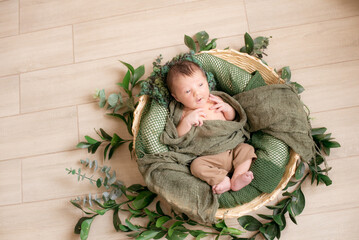 Cute little baby in a wicker basket decorated with green branches in a green knitted blanket at home on a light wooden background 
