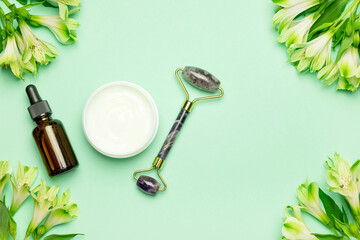 Face roller with cosmetic serum and face cream on a green background with flowers. Facial massage kit for lifting massage.