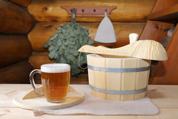 On a wooden desk a mug of light beer stands on a wooden lid near the accessories for the sauna on the background a log wall with hanging on it a bath broom and a hat.