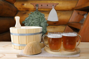 On a wooden desk a couple glass mugs of light beer stand on a wooden lid near the accessories for the sauna on the background a log wall with hanging on it a bath broom and a hat.