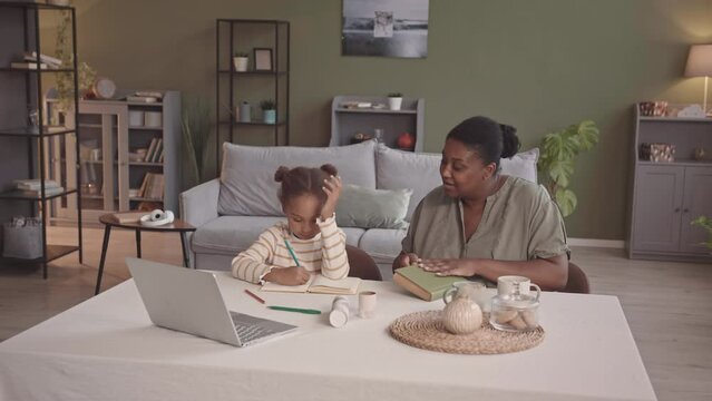 Stab slowmo of loving African-American mother and little daughter spending leisure time together at cozy home. Girl drawing while her mom reading sitting at table in living room