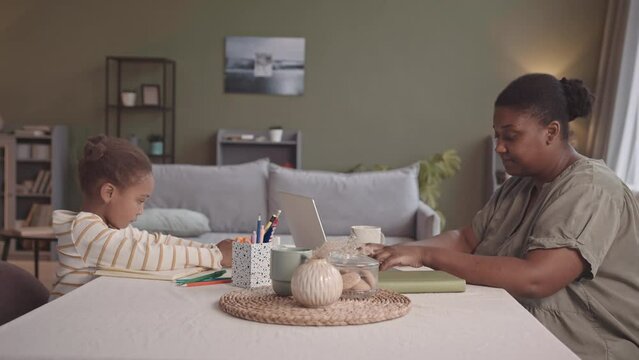 Medium slowmo of young body positive African-American woman working on laptop from home while her 5 year old daughter drawing sitting at desk in front of her in cozy living room