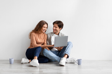 Satisfied young caucasian couple look at laptop planning new interior, sit on floor in empty room