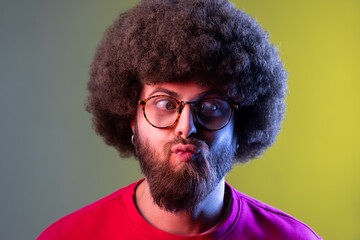 Portrait of crazy hipster man with Afro hairstyle standing with crossed eyes and looking with funny comedian face, wearing red sweatshirt. Indoor studio shot isolated on colorful neon light background