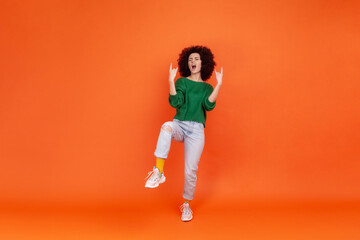 Fototapeta na wymiar Full length woman with Afro hairstyle wearing green casual style sweater showing rock and roll gesture, standing on one leg, yelling. Indoor studio shot isolated on orange background.