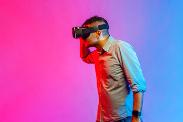 Side view of handsome man in shirt wearing virtual reality glasses, looking attentively aside, watching 3d video. Indoor studio shot isolated on colorful neon light background.