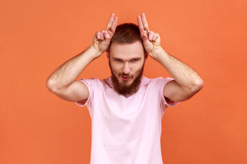 Fototapeta na wymiar Portrait of angry bully bearded man showing bull horn gesture with fingers over head, looking hostile and threatening, wearing pink T-shirt. Indoor studio shot isolated on orange background.