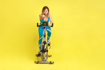 Full length portrait of blonde young adult sporty woman having cardio workout on exercise bike, pointing finger to camera, inviting you doing sport. Indoor studio shot isolated on yellow background.