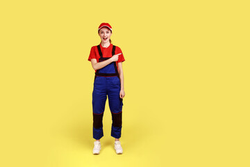 Full length portrait of excited amazed handy woman standing and pointing finger away, showing copy space for advertisement, wearing overalls and cap. Indoor studio shot isolated on yellow background.