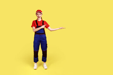 Full length portrait of amazed worker woman presenting copy space for advertisement on palm, looks at camera with excitement, wears overalls and cap. Indoor studio shot isolated on yellow background