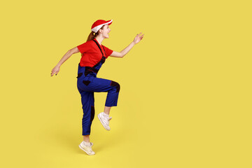 Side view of positive worker woman standing on one leg with raised arms, looking ahead, marching, happy expression, wearing overalls and red cap. Indoor studio shot isolated on yellow background