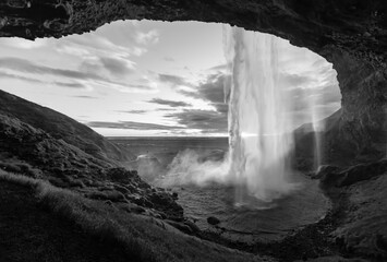 Seljalandsfoss magical Waterfall in the south of Iceland with Midnight Sun in Midsummer night with wonderful water curtain and cloudy sky seen from a cave behind the cascade, black and white greyscale