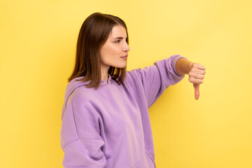 Side view of upset dissatisfied woman standing with thumb down, negative feedback, demonstrating disapproval gesture, wearing purple hoodie. Indoor studio shot isolated on yellow background.