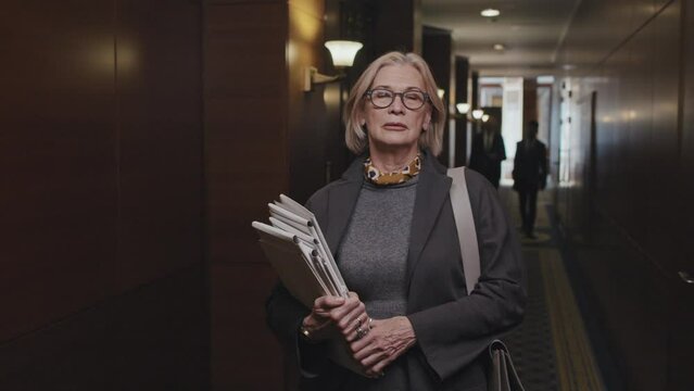 Waist up shot of senior businesswoman walking in corridor in office center with file folders