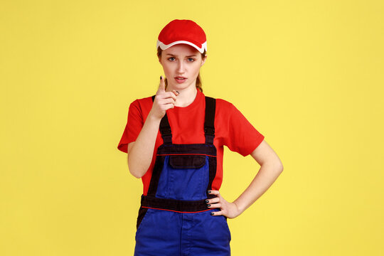 Portrait Of Bossy Confident Worker Woman Standing, Raising Finger, Showing Warning Gesture, Looking At Camera, Wearing Overalls And Red Cap. Indoor Studio Shot Isolated On Yellow Background.