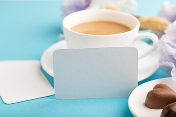 White business card with cup of cioffee, chocolate candies and iris flowers on blue pastel background. side view, copy space.