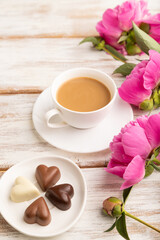 Cup of cioffee with chocolate candies, pink peony flowers on white wooden background. side view, close up.