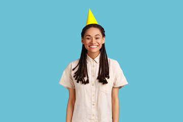 Portrait of optimistic pretty woman in party cone hat, looking at camera with toothy smile, rejoicing holiday celebration, wearing white shirt. Indoor studio shot isolated on blue background.