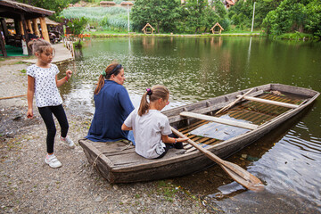 Family fun on old wooden boat at lakeshore. Mother with two daughters have leisure time by water. Summer day in nature.