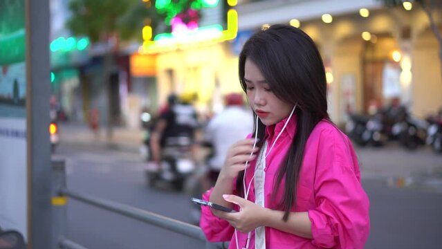 a cute Vietnamese girl waiting at a bus station is pulling her earphone to listen to her favorite song and sing along with it