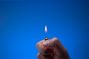 Lit small lighter with flame held between fingers by man. Close up studio shot, isolated on blue background