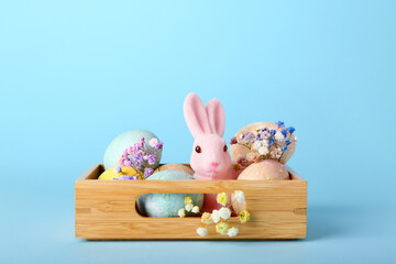 Box with beautiful Easter bunny, painted eggs and flowers on blue background