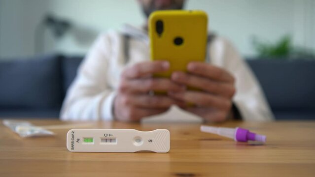 Caucasian man using a smart phone after makes a rapid home test for Covid19 virus with a positive result. Person is typing phone on wood table with antigen cassette of express test coronavirus