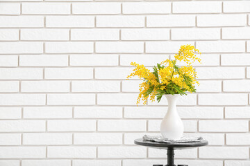 Fototapeta na wymiar Vase with beautiful mimosa branches on table near light brick wall in room