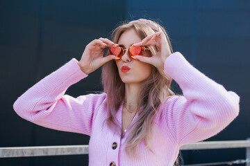 Lady chews gum in the shape of a heart. Beautiful girl in a pink sweater and jeans and pink glasses. Modern style, positive attitude, urban look