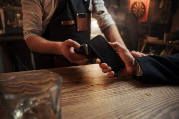 Caucasian male standing at bar tapping cellular device on card machine paying for drinks