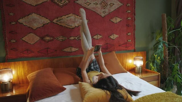 girl lying on the bed wearing parisian stockings or winter long socks on legs stretched to the wall keeping arms raised using mobile phone