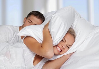 Despaired millennial wife on bed covers ears with pillow and suffers from snoring of sleeping husband