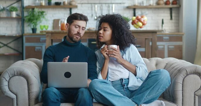 Happy husband and wife relaxing sitting on sofa enjoying using laptop computer on sofa, man looking at screen browsing internet, woman drinking tea, resting together in apartment