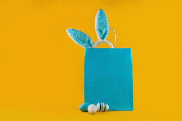 Blue shopping bag, rabbit ears and Easter eggs on a yellow background. Online shopping for the holidays. Symbols, traditions concept. Trending color of the year for Easter.