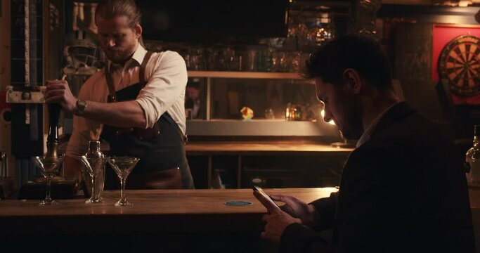 Caucasian male bartender making drinks for customer texting on cellular device 