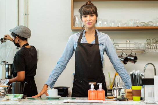 Successful small business owner in casual wearing apron standing at coffee shop.Portrait of restaurant owner barittra standing at counters and cafés .In the back, there is a waitress making coffee.
