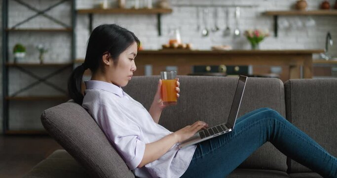 Asian woman drinking juice resting on couch using laptop looking at screen typing message at living room, young female chatting on computer browsing internet working online on sofa at home