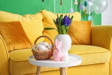 Flowers, basket of Easter eggs and rabbit on table near comfortable sofa