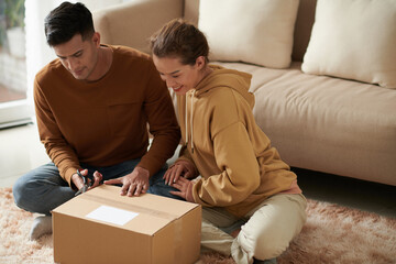 Young man opening the parcel with scissors with excited woman sitting near by him, they ordering...
