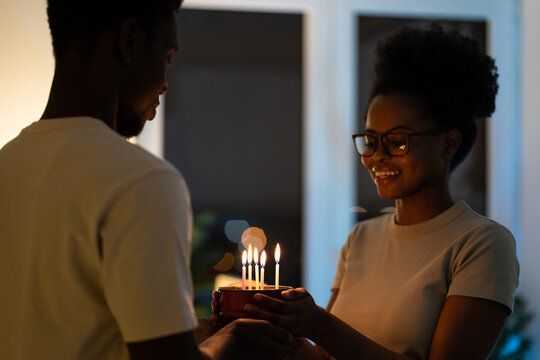 Celebration of love. Young happy african american couple holding small homemade cake with burning candles, celebrating special day wedding anniversary together at home in cozy romantic atmosphere.