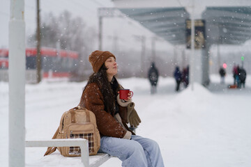 Tired but satisfied Asian traveler woman sitting on bench on snow-covered platform with mug of hot...